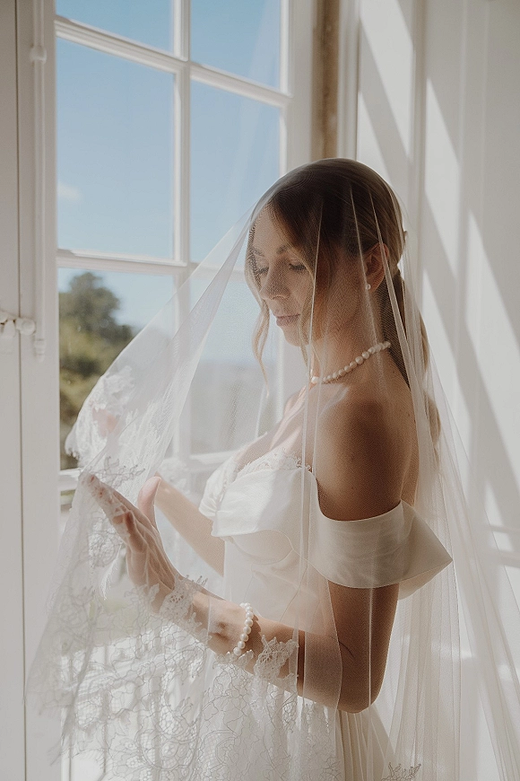 Bridal portrait of a bride with veil adjusting lace veil over her face by a bright window, wearing pearls and off-shoulder dress