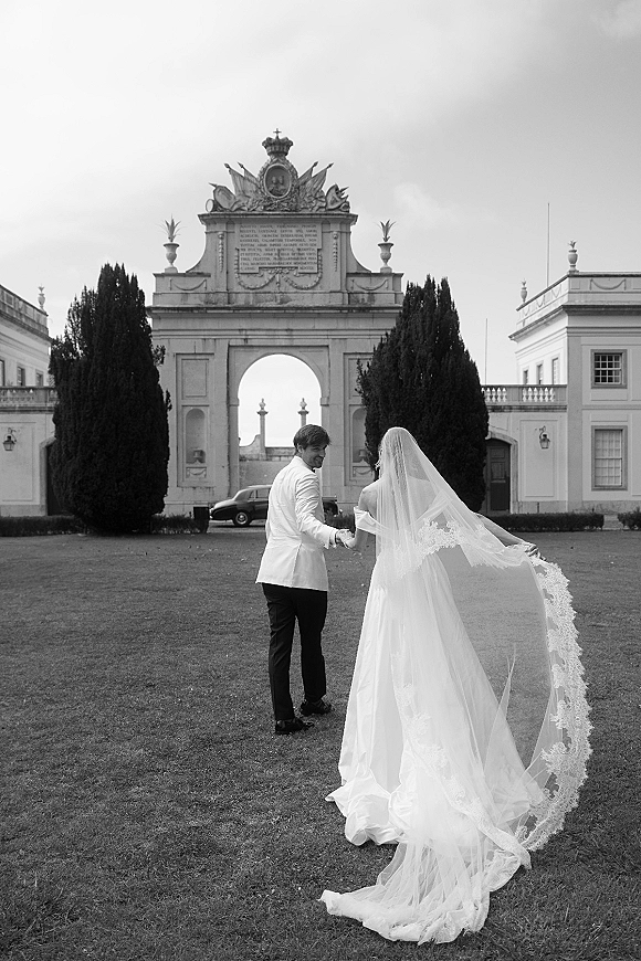 Wedding couple portrait of bride and groom walking away hand in hand, her long lace veil trailing beneath a stone palace archway