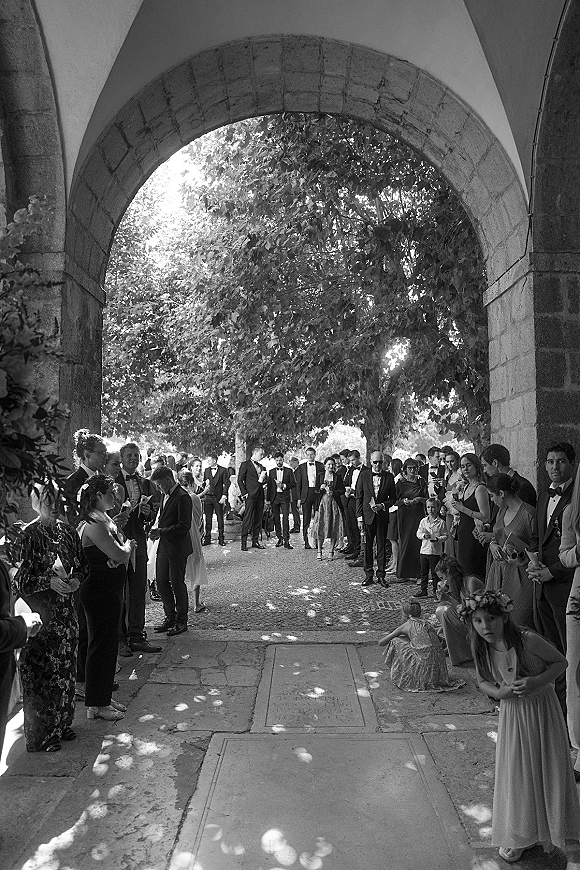 Wedding guests in formal attire line the aisle, with tuxedos and sunglasses under a stone archway on a cobblestone walkway