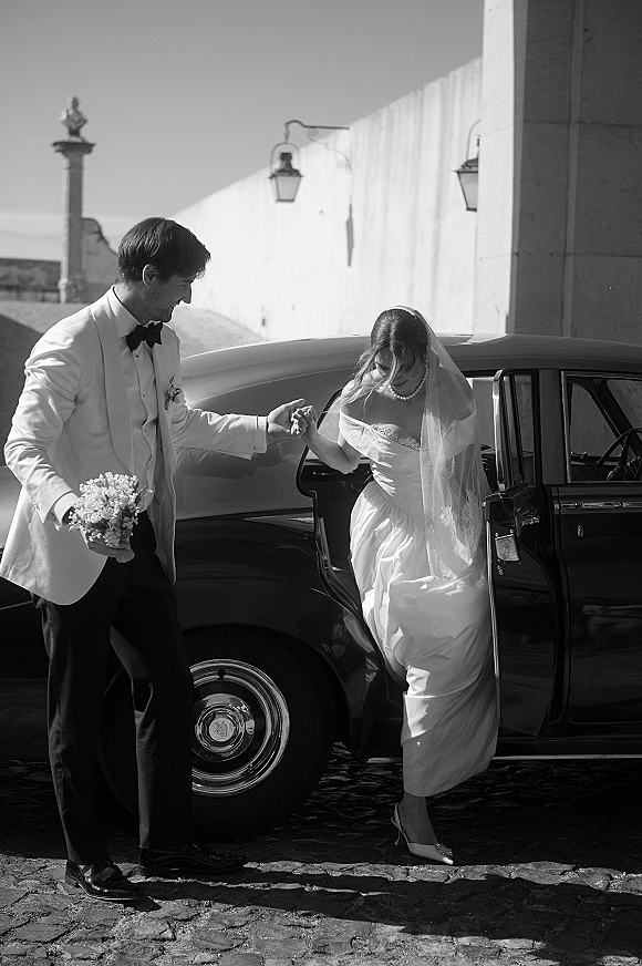 Wedding exit as groom in white dinner jacket helps bride in strapless gown into a vintage car, veil and bouquet in cobblestone courtyard
