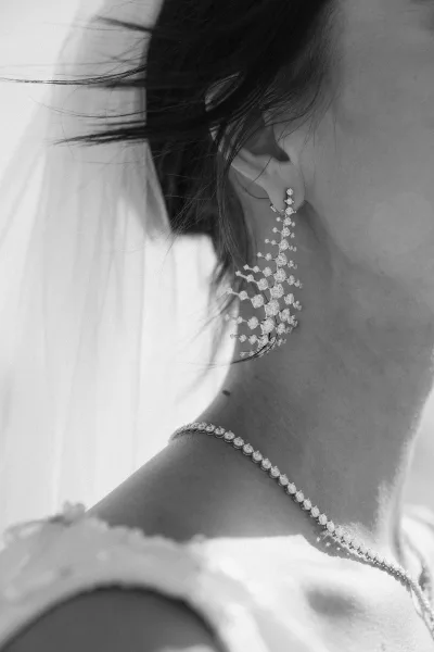 Bridal earrings sparkle as statement drop earrings beside a sheer veil, shown close up on the bride’s ear, neck, and shoulder
