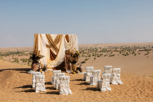Ceremony setup for a desert wedding ceremony with draped fabric backdrop, grounded florals and white chair covers on sand dunes under blue sky