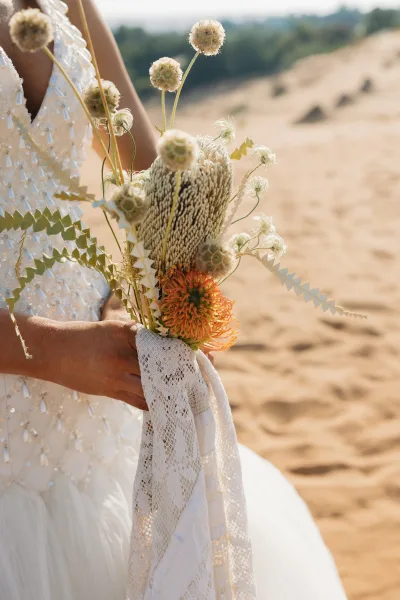 Bridal bouquet of dried flowers and wildflowers with orange pincushion and billy balls, lace wrap against sand dunes and sky