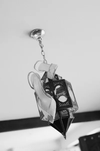 Bridal shoes, white high heel sandals with ankle straps, hang from a chain lantern fixture against the ceiling and wall backdrop