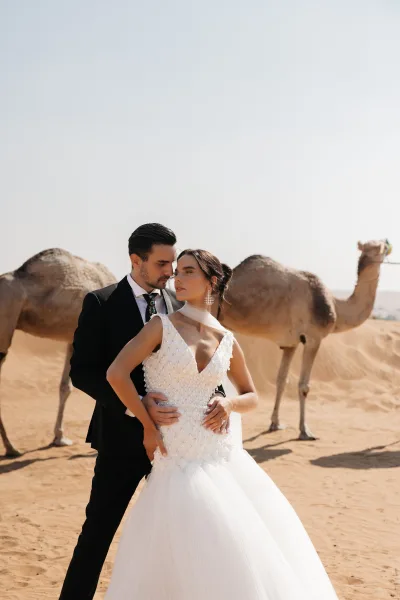 Couple portrait in desert wedding photos, groom holding bride from behind as camels stand on sand dunes under open sky