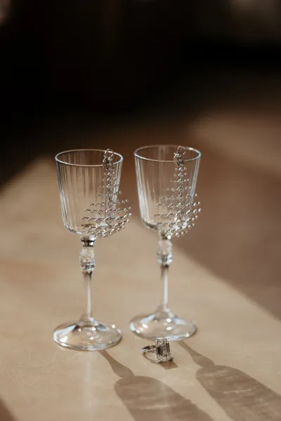 Wedding jewelry flatlay with bridal earrings and an emerald cut engagement ring beside wine glasses on a tabletop in warm shadows