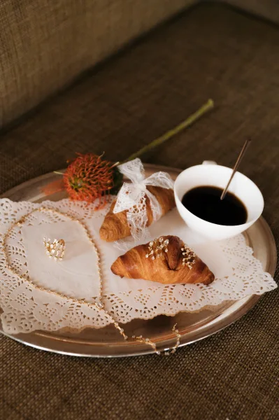 Bridal breakfast tray with wedding morning breakfast of croissants, coffee, lace ribbon, hair pins, and jewelry on an upholstered chair