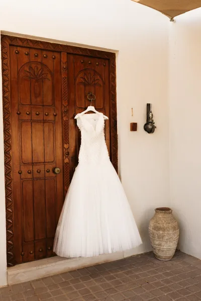 Wedding dress with beaded wedding dress bodice and tulle skirt hanging on a carved wooden door by a wall sconce on white stucco wall