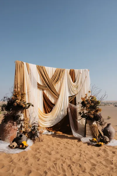 Wedding ceremony backdrop with draped wedding backdrop fabric, dried florals and ceramic urns set against sand dunes under blue sky