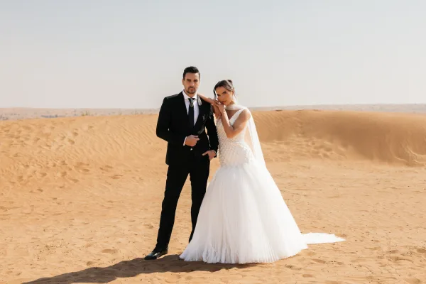 Couple portrait of bride in a wedding dress and veil holding groom in black suit on sand dunes under open desert sky