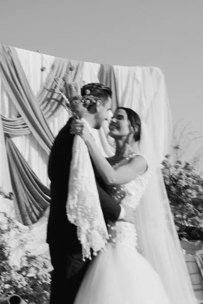 Wedding kiss portrait of bride and groom kissing, her veil and bouquet against a draped fabric backdrop with greenery indoors