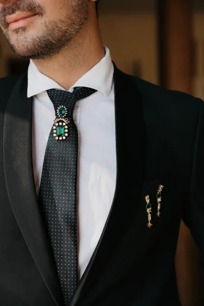 Groom accessories featuring a jeweled tie pin with emerald crystals on a black polka dot tie, black tuxedo and lapel pins against a warm neutral wall