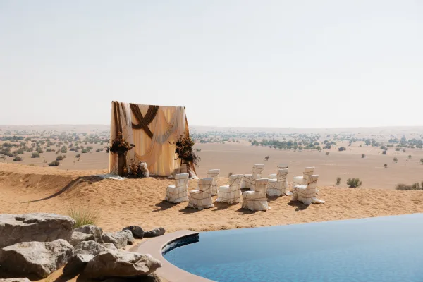 Ceremony setup with a draped fabric backdrop and floral arrangements at a desert wedding ceremony, with sand dunes and a pool behind chairs with sashes