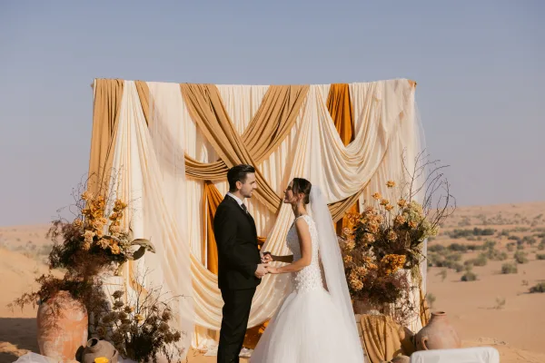 Ceremony moment with bride and groom holding hands beneath a draped fabric arch, roses and clay vases against desert dunes backdrop