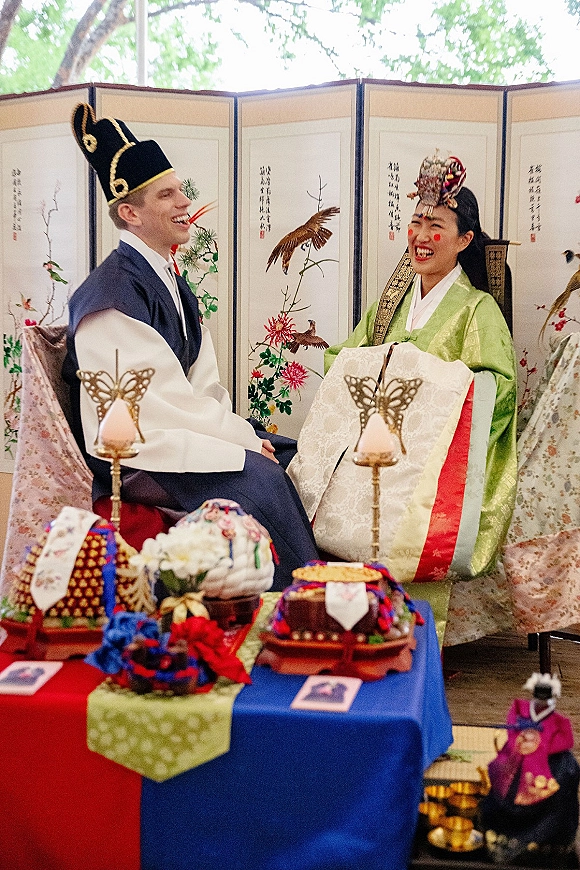 Wedding ceremony moment with bride and groom in hanbok smiling beside an offering table, lanterns, and folding screen under trees