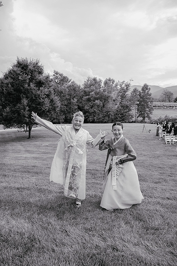 Wedding guest portrait of a smiling woman in a hanbok, wearing glasses, standing on a lawn with ceremony chairs and mountains behind