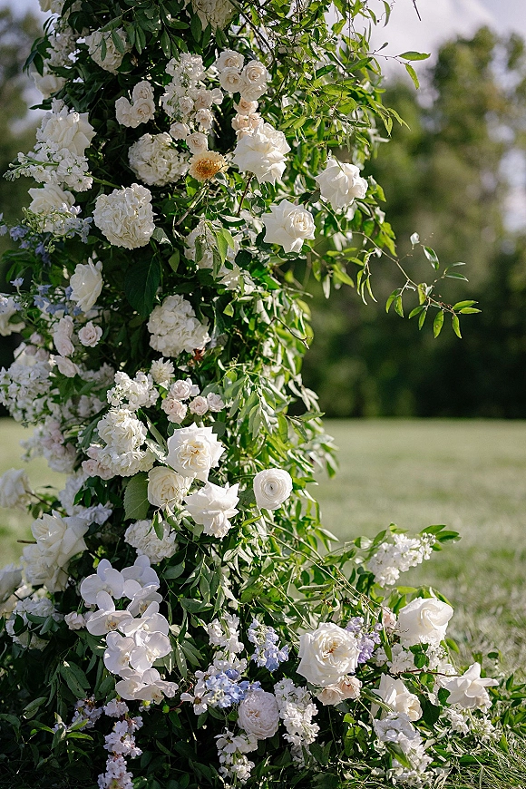 Ceremony floral arch with white roses and greenery, dotted with hydrangeas and light blue blooms on a sunlit lawn with trees