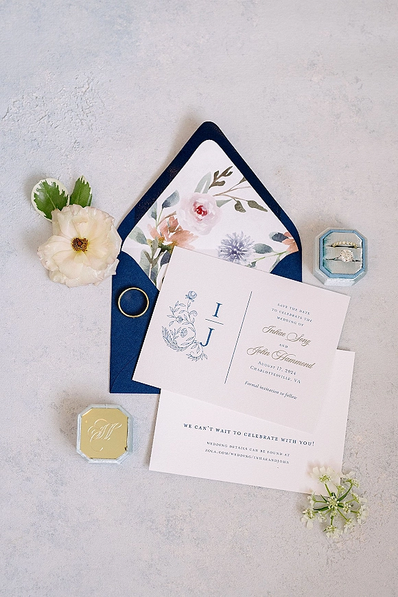 Invitation suite wedding invitation flat lay with navy envelope, floral liner, gold wax seal, ring box and flowers on light gray stone surface