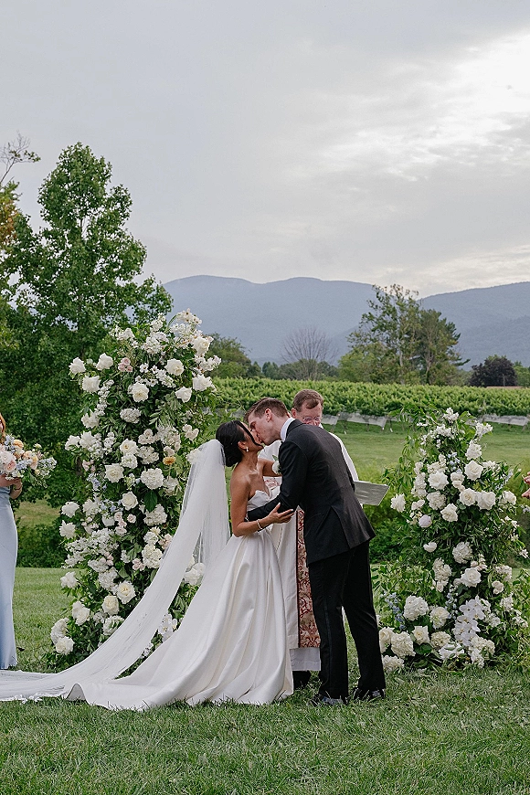 Wedding kiss beneath a white floral arch as the bride’s long veil drapes behind her and the groom in a tux, with vineyard rows and mountains beyond