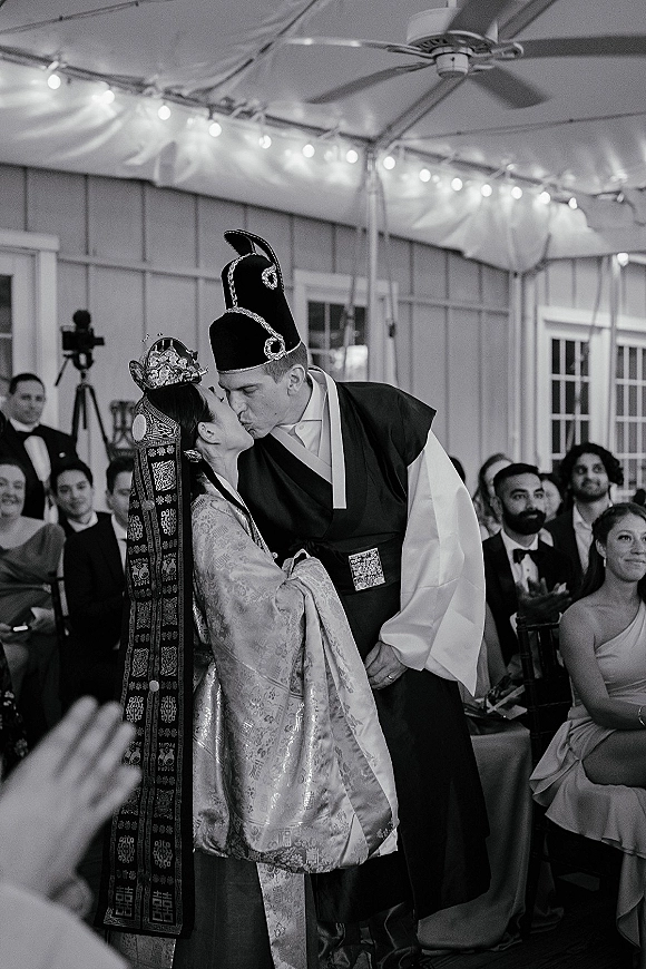 Wedding kiss in traditional Korean wedding robes as guests watch under a tent canopy with string lights and windows behind