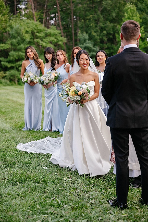 Ceremony moment as bride walks down the aisle smiling at groom, holding a bouquet with cathedral veil flowing on a garden lawn