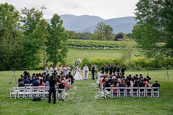 Outdoor wedding ceremony with a white floral arch and petal-strewn aisle, guests seated in a vineyard with mountain views under clouds