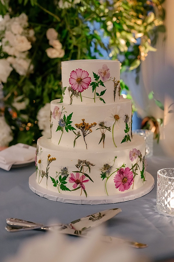 Wedding cake with pressed flower wedding cake design on a table, three tiers of white buttercream with botanical greenery against a soft-lit backdrop