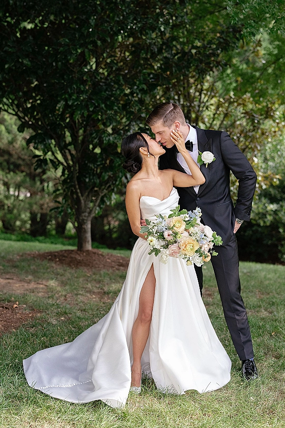 Wedding couple portrait of bride and groom kiss, bride touching his face, holding pastel bouquet in a garden with trees and lawn