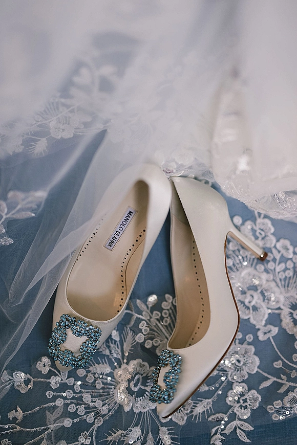 Wedding shoes in a flat lay, white high heel pumps with blue crystal buckles on lace and tulle veil over a blue surface