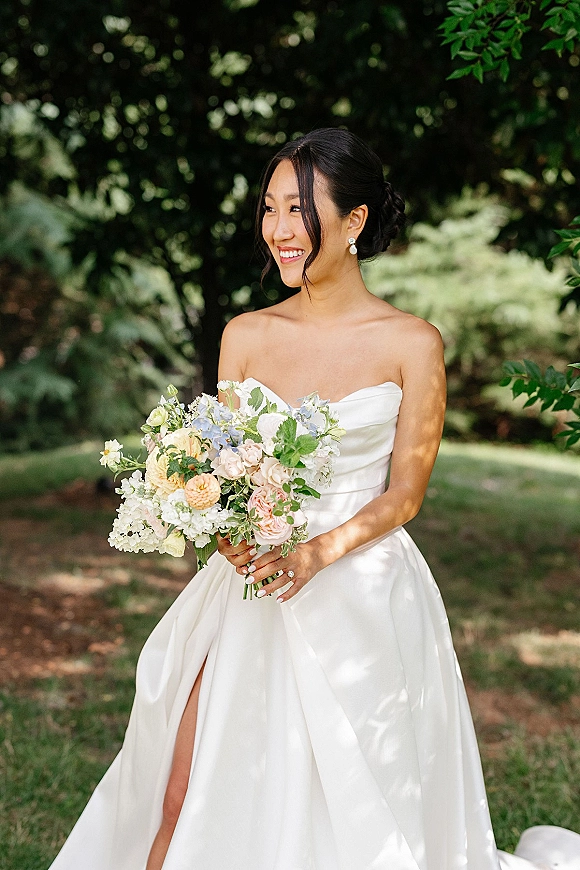 Bridal portrait of a bride holding bouquet in a strapless wedding dress, smiling in a lush garden with trees and lawn behind her