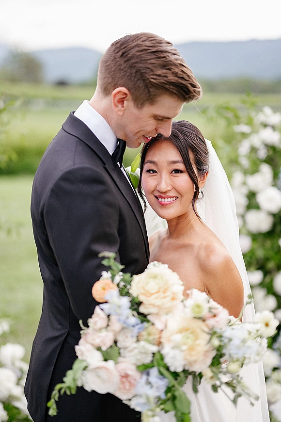 Couple portrait of bride and groom embrace as he kisses her forehead, bride holding bouquet, with floral arch and mountains behind