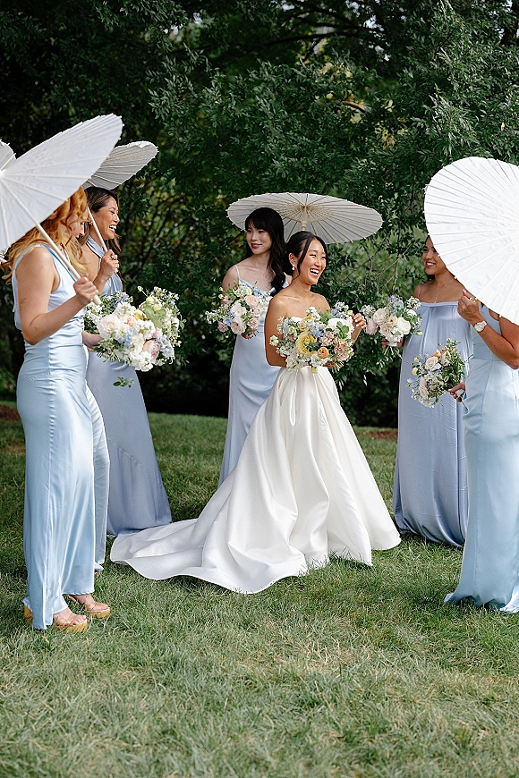 Bride with bridesmaids in a bridal party portrait, holding bouquets and white parasols on a grassy lawn with green trees behind
