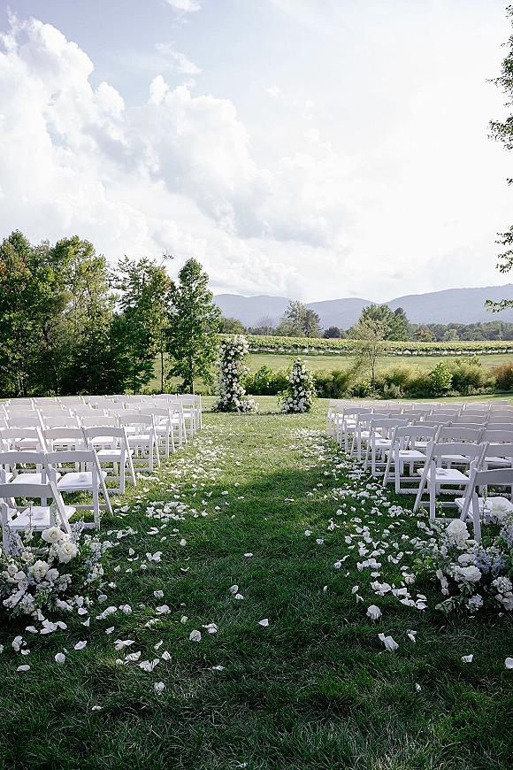 Outdoor ceremony setup with white folding chairs and white rose petal aisle, framed by vineyard rows and mountains under a cloudy sky