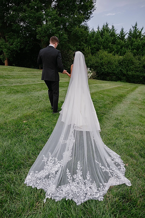 Wedding couple portrait of bride and groom walking away hand in hand on a lawn, her long veil trailing behind under cloudy sky