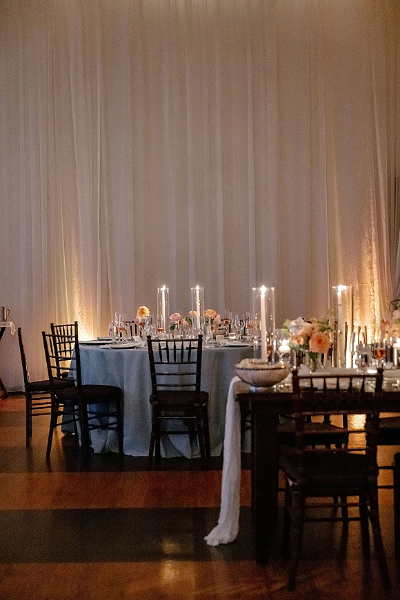 Reception tablescape with round wedding table decor, tall cylinder candles and floral centerpieces, set before a white drape backdrop with uplighting