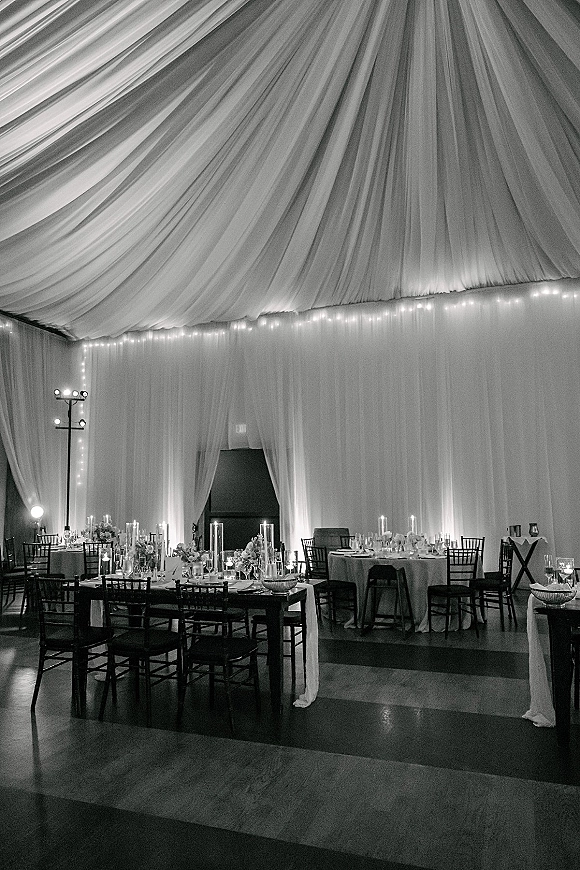 Reception decor under wedding tent draping with string lights, ceiling fabric swags, candlelit centerpieces, and a dance floor framed by uplighting