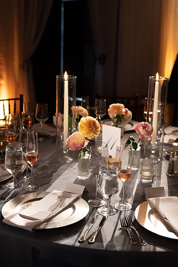 Reception tablescape with a candlelit wedding table featuring tall taper candles, peach blooms, menus and place cards in a dark, draped room