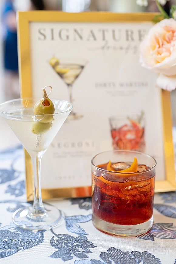 Signature cocktail sign in a gold frame showing martini and rocks glass drinks with olive and orange peel, set beside florals on tablecloth