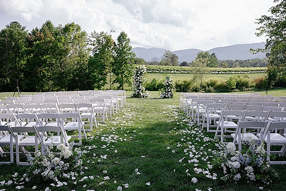 Ceremony setup with outdoor ceremony seating, white folding chairs framing a rose petal aisle and floral columns on a vineyard lawn with mountains