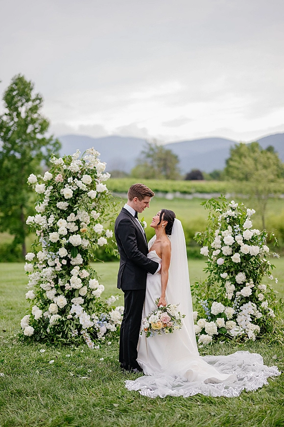 Couple portrait of bride and groom portrait kissing under a floral arch, her veil and bouquet flowing on a mountain lawn backdrop
