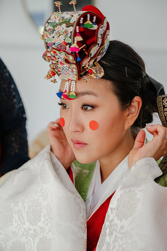 Bride portrait wearing a traditional bridal headdress with pearl tassels and cheek makeup dots, with helper hands adjusting pins indoors