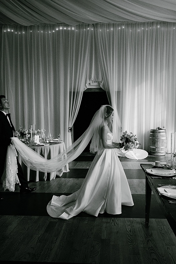 Bride portrait with a long veil and bouquet, glancing over her shoulder at an indoor reception entrance with string lights and taper candles