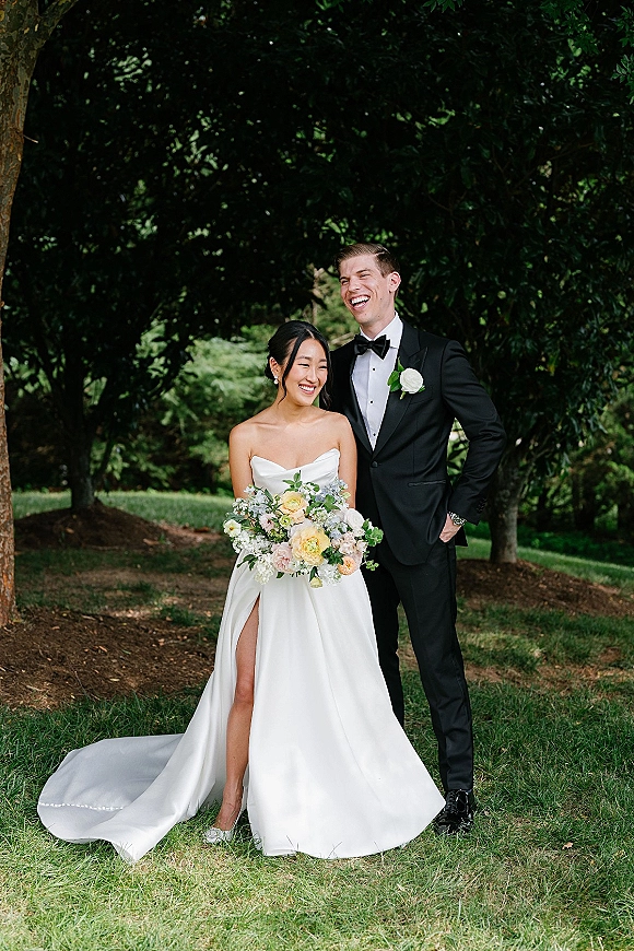Couple portrait of bride and groom laughing, bride holding a rose bouquet in a strapless satin wedding dress on a shaded lawn
