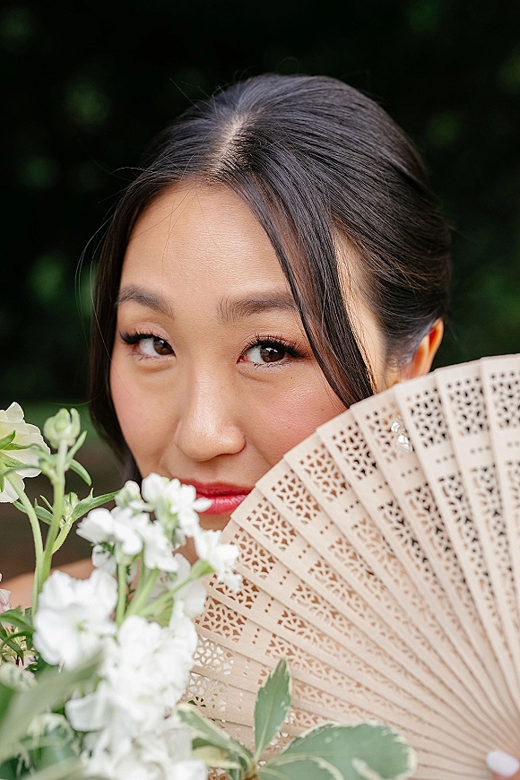 Bridal portrait of bride holding fan, a lace hand fan partly covering her face, with red lipstick and bouquet against dark foliage background