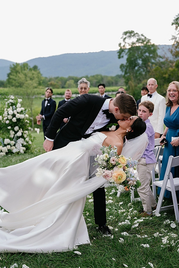 Wedding kiss moment as groom dips bride in strapless dress holding a pastel bouquet, long veil flowing on an outdoor lawn with mountains behind