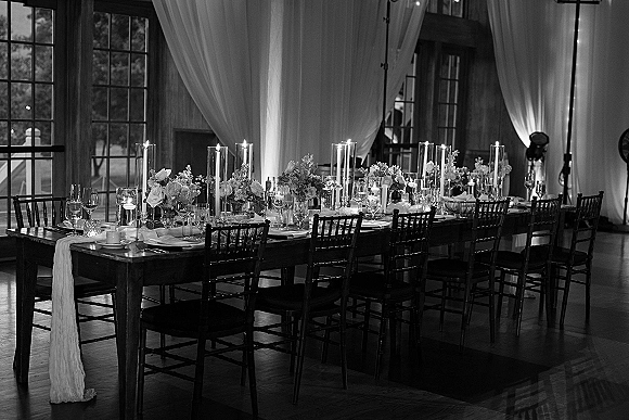 Reception tablescape on a long banquet table wedding setup with taper candles, glass cylinders, floral centerpieces, and chiavari chairs by large windows