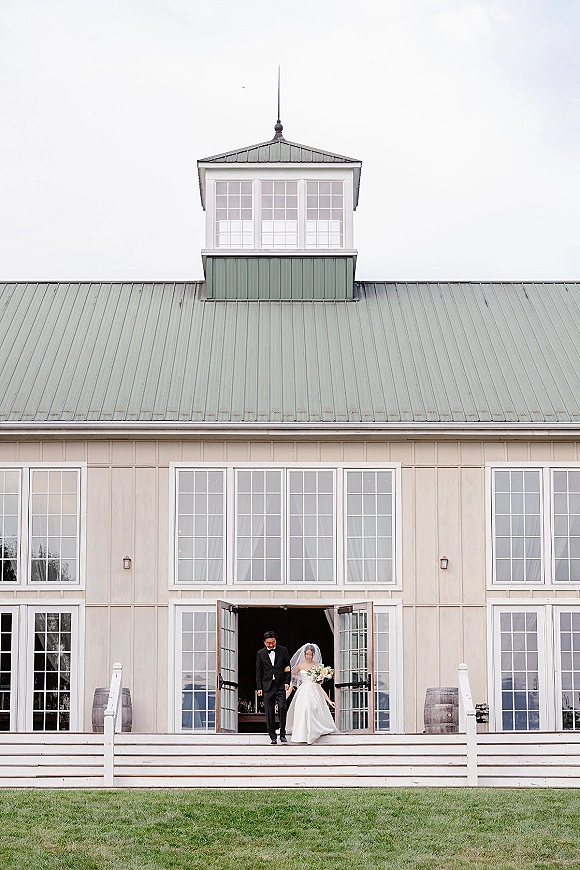 Wedding exit moment as bride and groom exit holding hands down barn steps, her long veil trailing, both smiling under overcast sky
