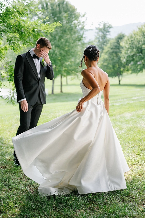 Wedding first look as bride in a strapless ball gown approaches the groom, his emotional reaction on a sunny lawn with trees and hills