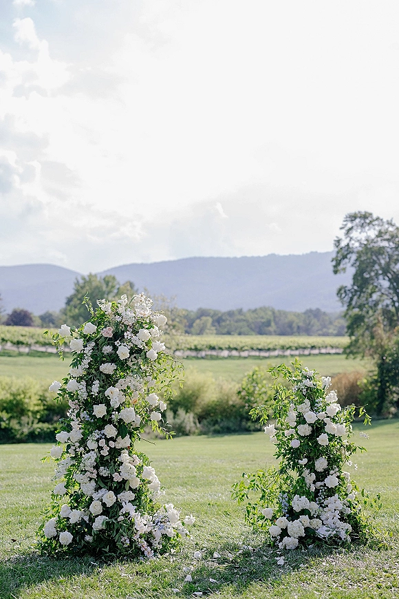 Wedding ceremony florals with floral ceremony pillars in white blooms and greenery on a grass lawn with vineyard rows and mountain backdrop