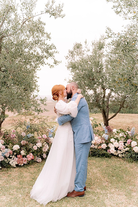 Wedding kiss as bride in a puff sleeve gown and updo embraces groom in a light blue suit amid roses in an olive grove on rolling hills